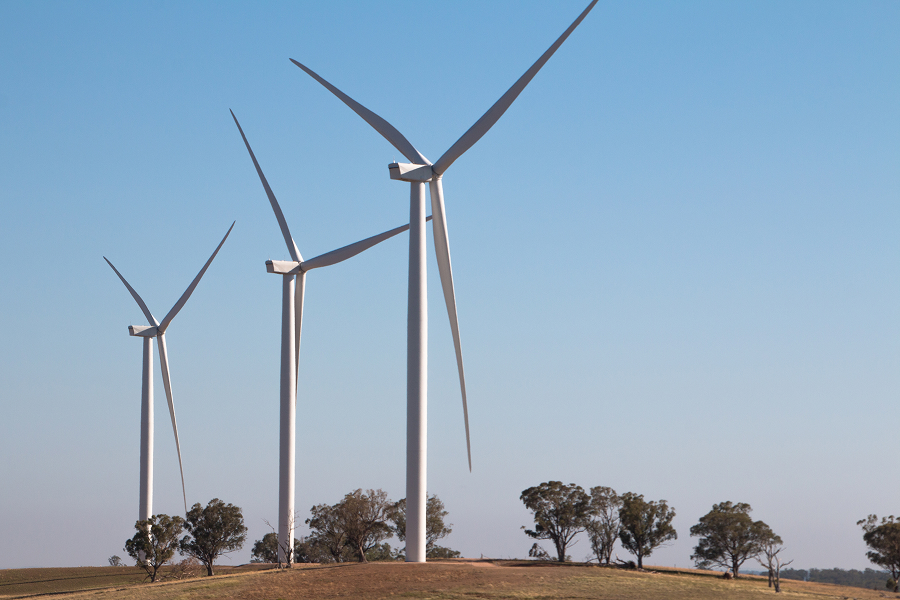 Imagen de tres aerogeneradores en un campo, con árboles al fondo y un cielo despejado. Las turbinas están girando, aprovechando la energía del viento.