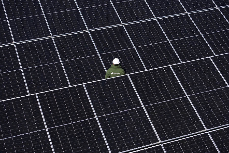 Trabajador con casco blanco y chaqueta verde caminando entre filas de paneles solares en una planta fotovoltaica.